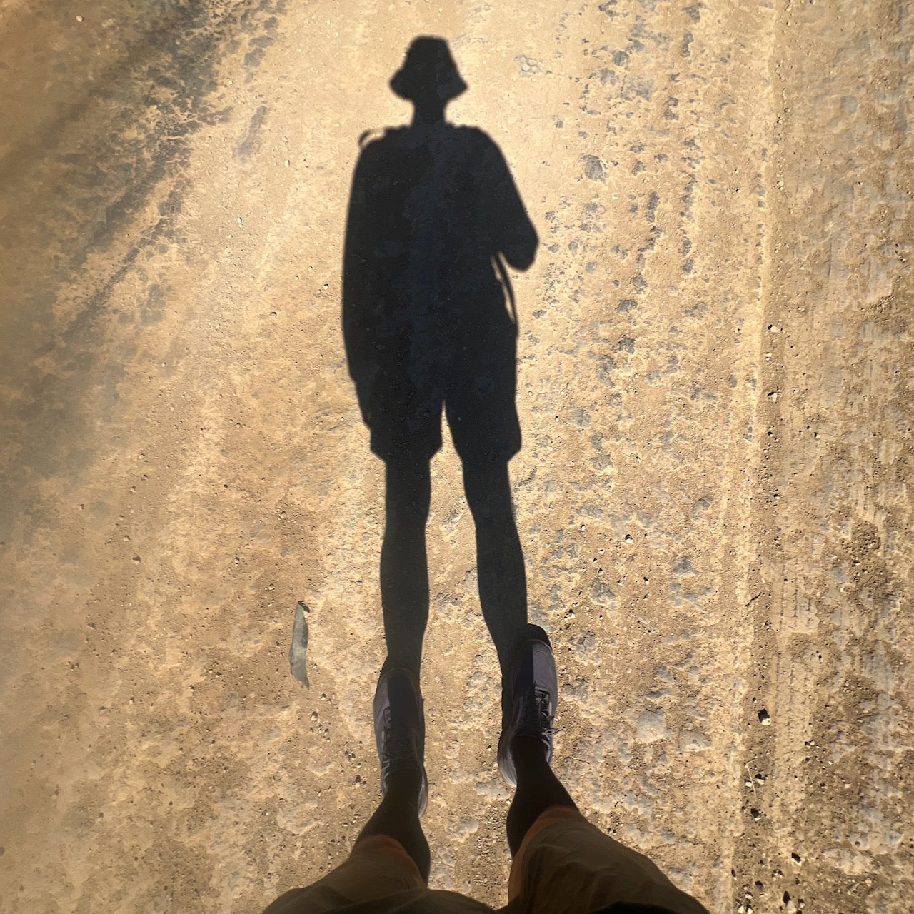 A person's shadow in a hat falls on a sunlit dirt road, their feet clad in Mackall - Forged Iron + Chiseled Stone + Gum all-terrain rucking shoes.
