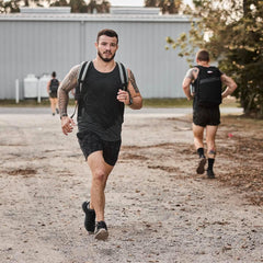 Athletic man with tattoos running outdoors wearing black tank top, shorts, backpack, and black GORUCK trainers