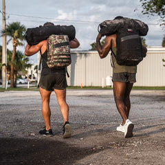Two muscular men carrying weighted duffel bags, wearing tactical GORUCK backpacks, walking outdoors