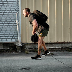 Man wearing GORUCK black ballistic trainers and backpack, outdoors on concrete next to metal siding
