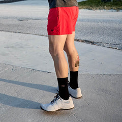 Man wearing red GORUCK ballistic training shorts, black socks, and white athletic shoes standing outdoors on concrete