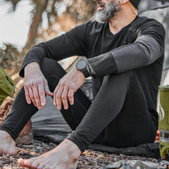Man wearing black merino wool base layer top and leggings sitting barefoot outdoors near camping gear