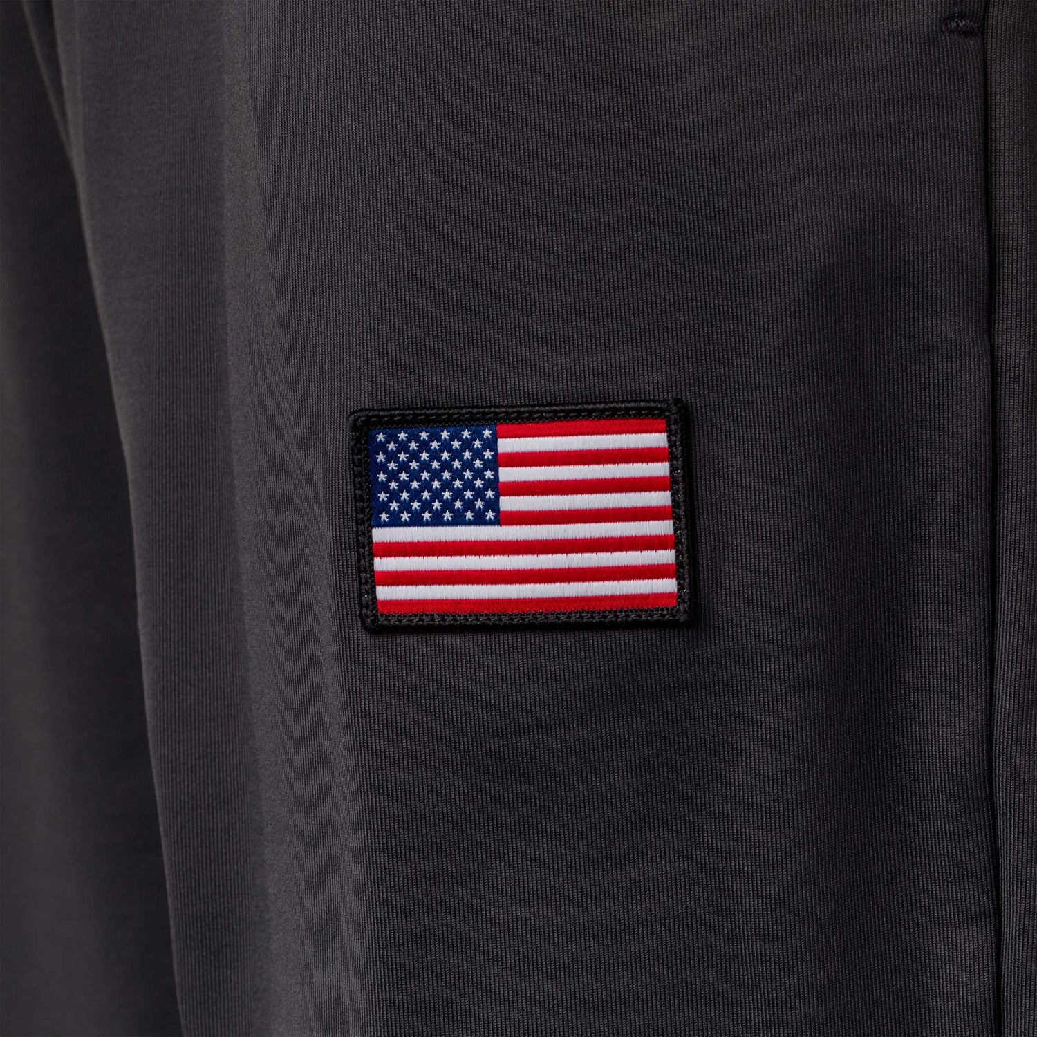 A small American flag patch is sewn onto dark fabric, likely part of the Men’s Training Performance Joggers.