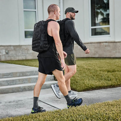 Two men walking on a sidewalk next to a grassy area, dressed in athletic gear with GORUCK Rough Runner backpacks in Midnight Frogskin and Acid Lime. One of the men wears a cap as they pass by a building with large windows in the background.