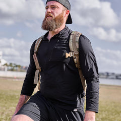 Bearded man in a black Men's USA Long Sleeve Performance Half Zip - ToughMesh and cap, carrying a khaki backpack outdoors on a cloudy day. Backed by the Scars Lifetime Guarantee for lasting durability.