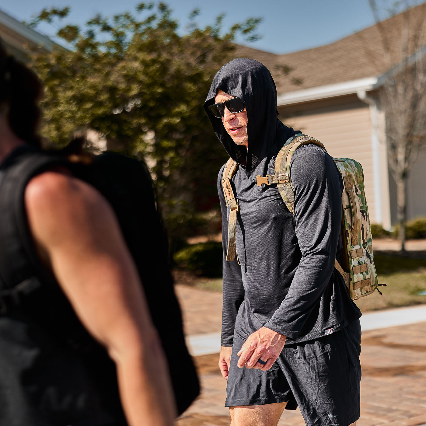 A man wears the Men's USA Sun Hoodie - ToughMesh, sunglasses, and shorts while walking outdoors with a camouflage backpack on a sunny day.