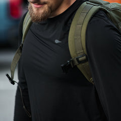 A person in a Men's Long Sleeve Performance Tee made of ToughMesh™ fabric and a green backpack is outdoors. Their partially visible face reveals a rugged beard.