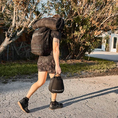 A person strides confidently outdoors, dressed in black athletic gear and Men's Ranger Panties - ToughStretch from GORUCK. With a large backpack slung over their shoulder and a smaller GORUCK bag in hand, they navigate the sunny path lined with trees and chain-link fences.