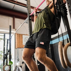 A person with a backpack performs a pull-up using a pink bar in a gym setting, wearing a green shirt and black Men’s USA Training Shorts by GORUCK, known for their ToughStretch durability and SCARS Lifetime Guarantee. Gymnastic rings and exercise equipment are visible in the background, emphasizing the rigorous workout environment.