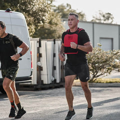 Two men are jogging outdoors on a sunny day, with one wearing a red weighted vest. Dressed in GORUCK Men’s USA Training Shorts featuring their ToughStretch fabric, they run past a white van and industrial buildings. Trees and a clear sky serve as the backdrop as they confidently enjoy the SCARS Lifetime Guarantee of their gear.