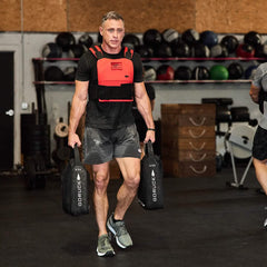 A man is working out in a gym, clad in GORUCK's Men’s USA Training Shorts - ToughStretch and sporting a weighted vest while holding two hefty sandbags. As he moves decisively through weights, boxes, and balls, his equipment boasts the SCARS Lifetime Guarantee. Another individual can be seen partially on the side.