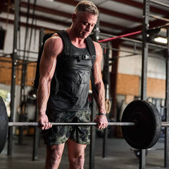 A man in a gym lifts a barbell while wearing a weighted backpack. Dressed in a black tank top and GORUCK Men’s USA Training Shorts made with ToughStretch fabric, he stands amid metal equipment as natural light filters through large windows.