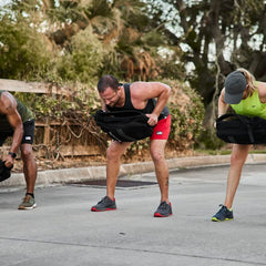Three people outdoors performing bent-over rucking exercises with black GORUCK weighted backpacks