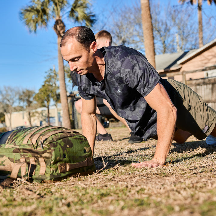 A man does push-ups outdoors on grass near a camo bag, wearing the Men’s USA Performance Tee - ToughMesh for quick-drying performance; another person is visible in the background.