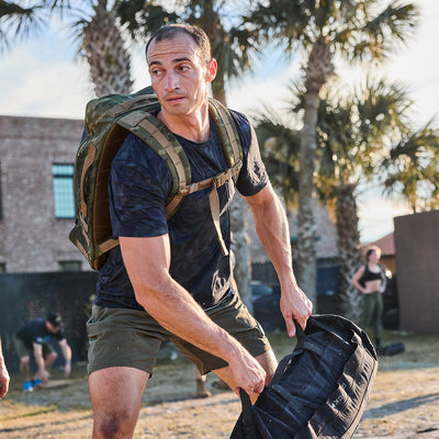 A man in athletic gear lifts a sandbag outdoors, wearing the Men’s USA Performance Tee - ToughMesh and a backpack, with palm trees in the background.