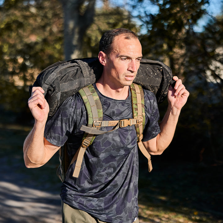A man carries a heavy sandbag outdoors in sunlight, wearing the Men’s USA Performance Tee - ToughMesh made from quick-drying, featherweight fabric.