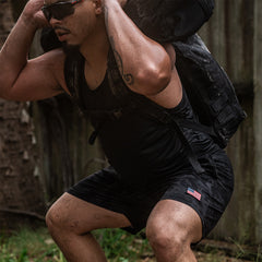 A man squats outdoors with a heavy sandbag on his shoulders, wearing Men’s USA Training Shorts - ToughStretch, featuring an American flag patch and backed by the SCARS Lifetime Guarantee.
