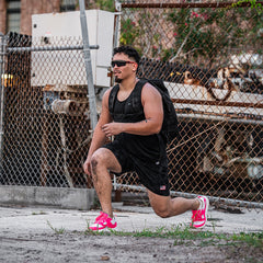 Man wearing black attire and bright pink sneakers lunges outdoors by a chain-link fence, showcasing Men’s USA Training Shorts - ToughStretch for durability and flexibility.