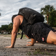 A person does push-ups outdoors on dirt, wearing a weighted backpack and Men’s USA Training Shorts - ToughStretch with a small US flag patch. These durable shorts are built to last and come with the SCARS Lifetime Guarantee.
