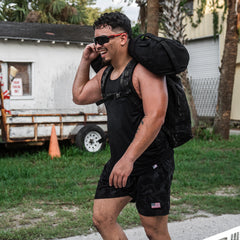 A smiling man wears Men’s USA Training Shorts - ToughStretch and workout gear, carrying a heavy backpack outdoors with sweat on his arms and sunglasses on.