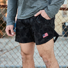 A person in Men’s USA Training Shorts - ToughStretch, featuring black camo fabric and a small U.S. flag patch, stands by a chain-link fence.
