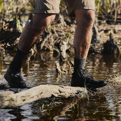 A person wearing black boots and beige shorts strides through a muddy swamp, stepping on a wet log. Their legs, splattered with mud and surrounded by water and reeds, are clad in GORUCK Merino Challenge Socks - Crew for comfort in every step.