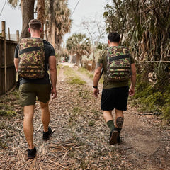 Two people walking on a dirt path surrounded by trees and a wooden fence, their attire hinting at adventure. Clad in green and black with camouflage backpacks, they tread through dry leaves, perhaps in a forest or rural area, with the comfort of GORUCK's Merino Challenge Socks - Crew cushioning each step.