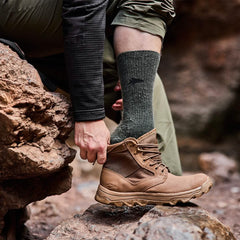 On a rocky trail, a person sitting on a stone adjusts their brown hiking boot, proudly Made in the USA. They are wearing GORUCK's Merino Challenge Socks - Crew in green and green cargo pants. A dark long-sleeve shirt is visible as well. The focus is on the boot and rugged terrain, equipped for any adventure with Scars Lifetime Guarantee.