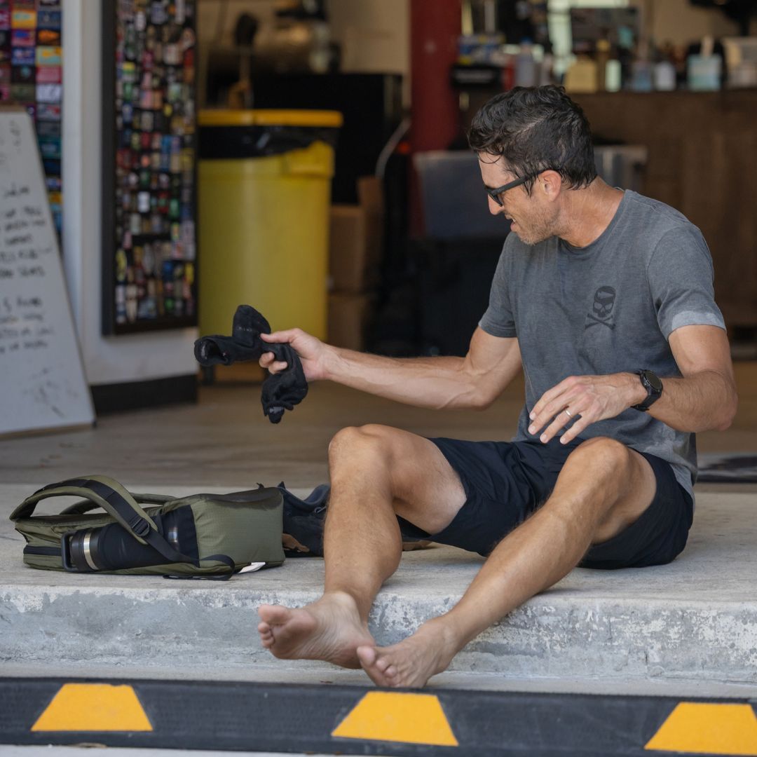 A barefoot man in sunglasses sits on a curb, holding a pair of Merino Challenge Socks - Ankle (No-Show), with a camera bag beside him.
