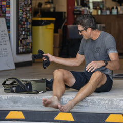A barefoot man in sunglasses sits on a curb, holding a pair of Merino Challenge Socks - Ankle (No-Show), with a camera bag beside him.