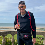 A man wearing the Men's Rucking Shirt - Merino Wool stands by a stone railing with sunglasses and a drink, ocean in the background.