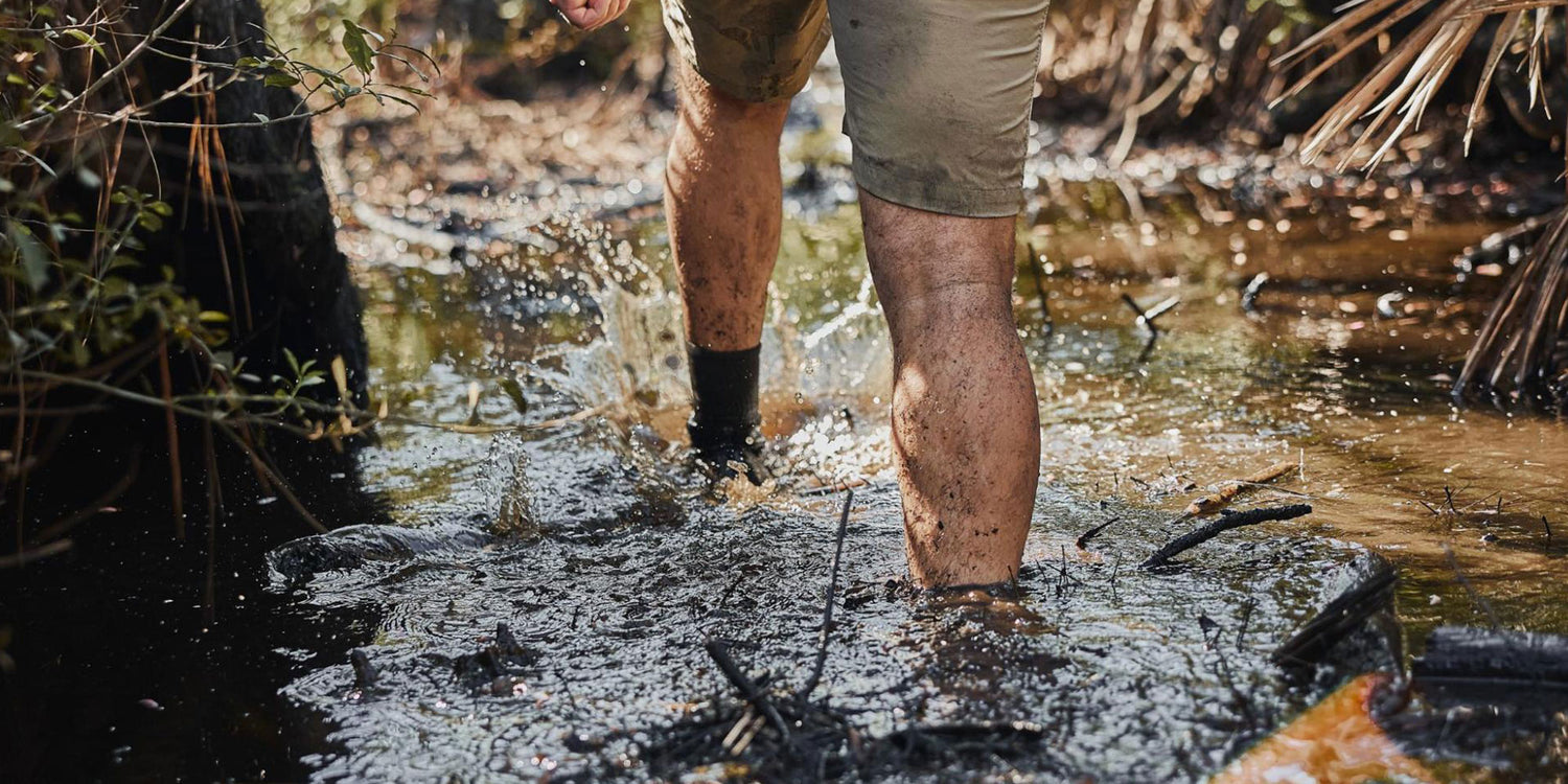 Person walking through muddy water in shorts, legs splashing and covered with mud, surrounded by plants.