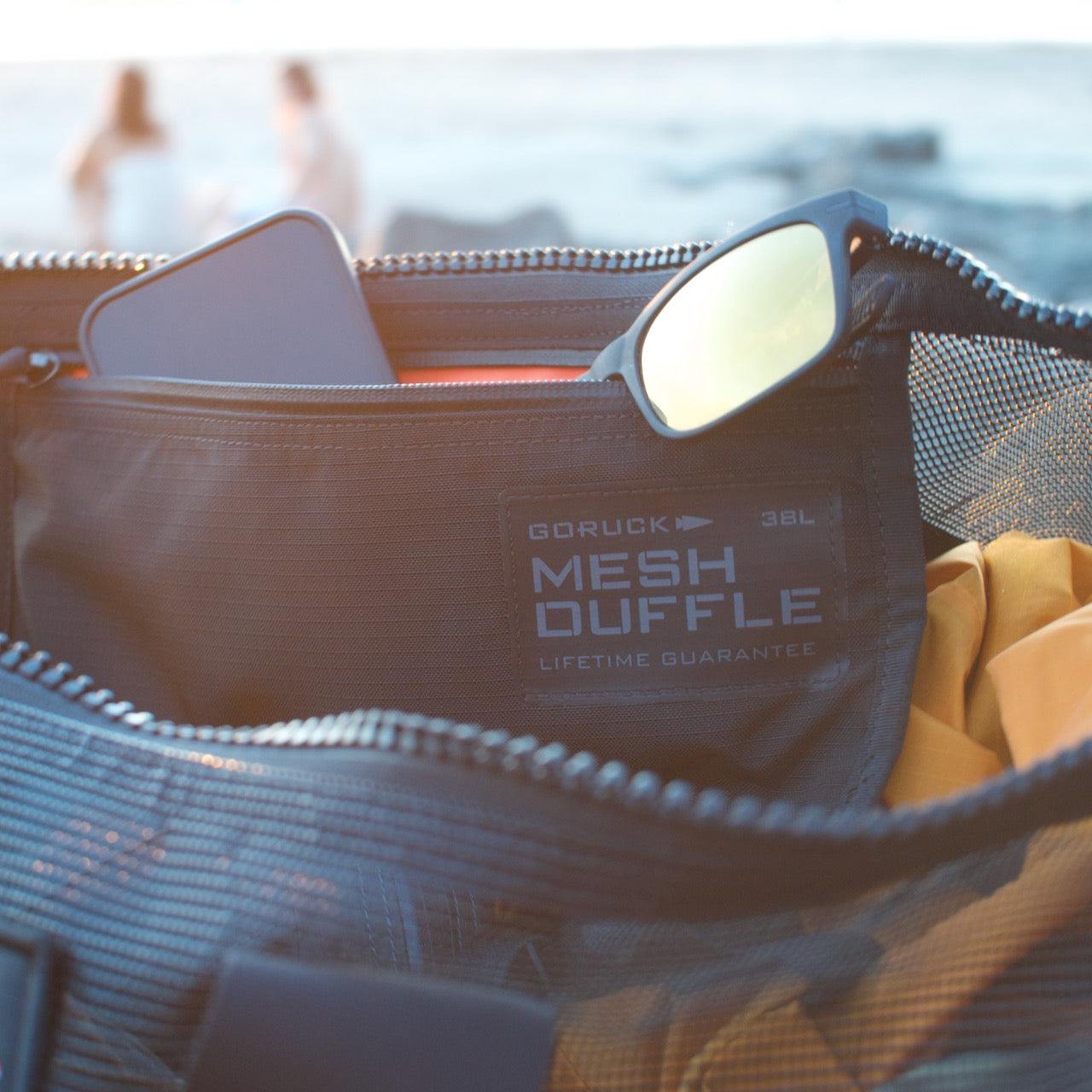 A Mesh Duffel Bag containing sunglasses and a smartphone sits on the beach, with two blurred people in the background.