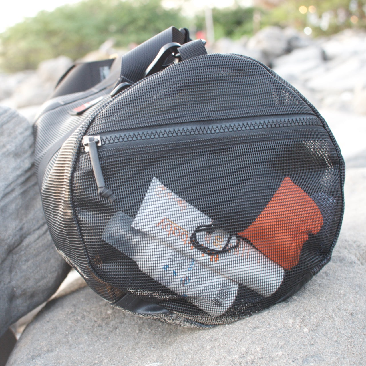 The Mesh Duffel Bag, shown in black with toiletries and clothing on outdoor rocks, is ideal for carrying wet clothes after a beach day.