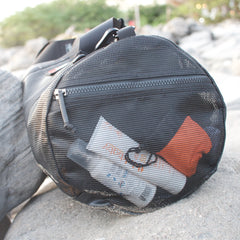 The Mesh Duffel Bag, shown in black with toiletries and clothing on outdoor rocks, is ideal for carrying wet clothes after a beach day.
