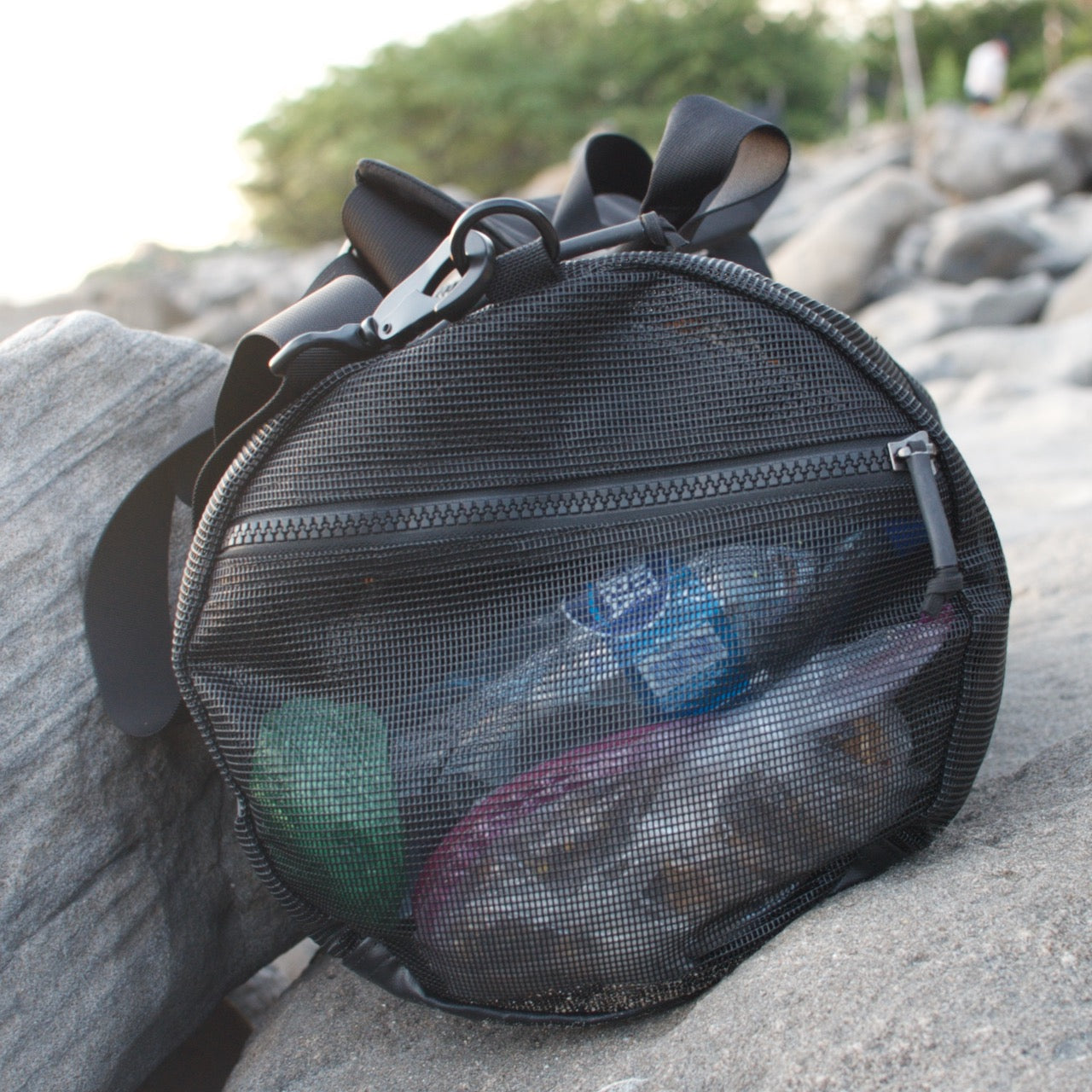 A Mesh Duffel Bag filled with plastic bottles and trash sits on outdoor rocks.