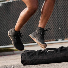 Person jumping in black GORUCK boots over a black rucking gear bag on pavement with chain-link fence background
