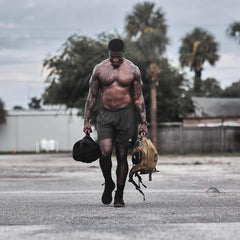 Muscular shirtless man walking outdoors carrying GORUCK rucking bags with palm trees in background