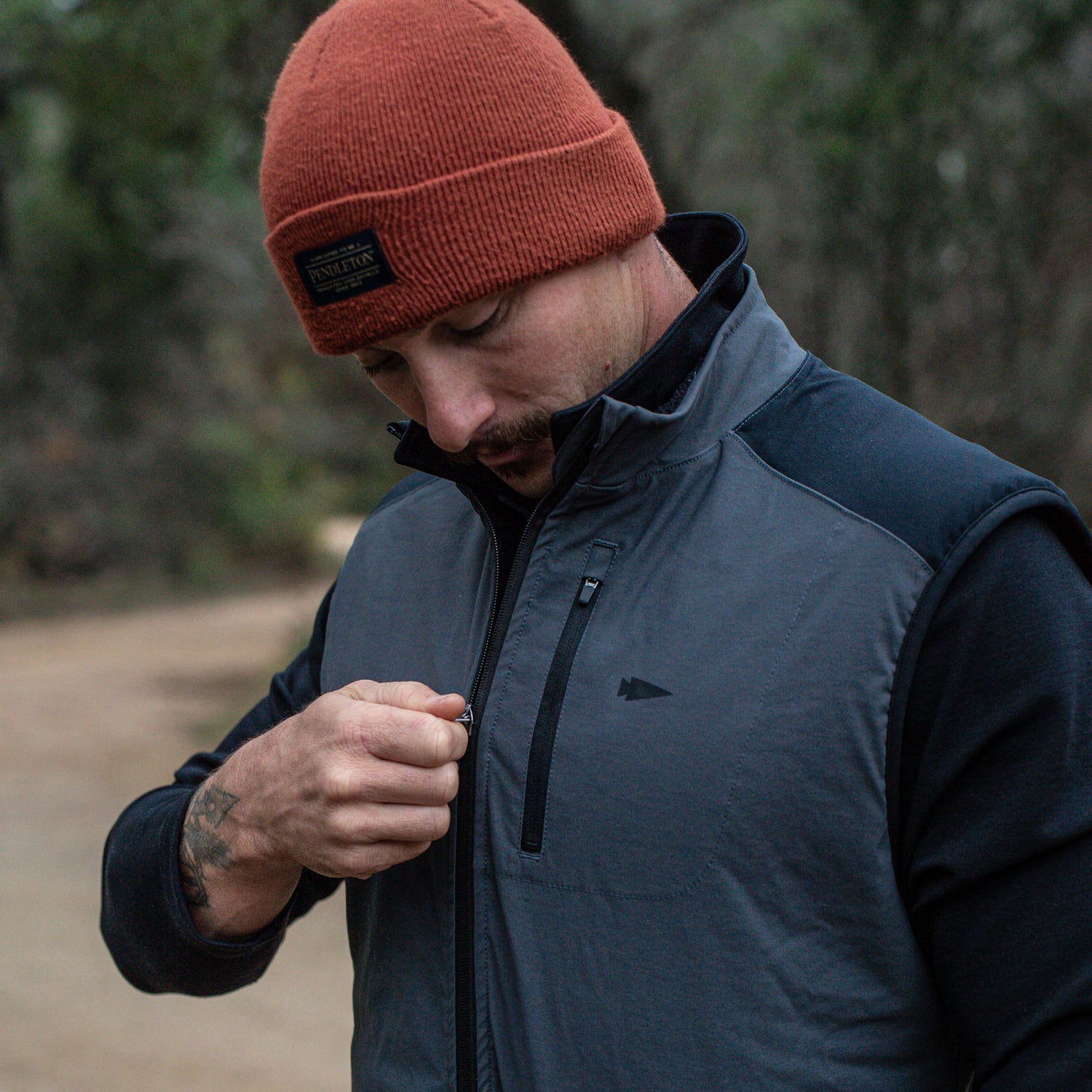 A man in a red beanie and jacket zips up his Men's Vest of Power - ToughDry® + Fleece outdoors, with blurred trees in the background.
