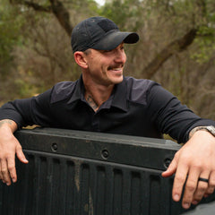 Man in black GORUCK cap and shirt leans on truck bed outdoors, smiling in wooded setting