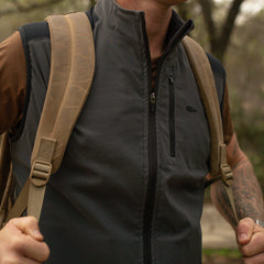 A man outdoors wears the Men's Vest of Power - ToughDry® + Fleece in gray and holds the straps of his tan backpack with both hands.