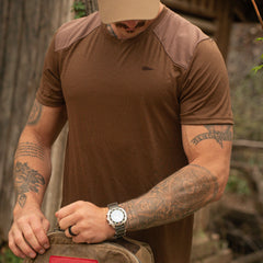 Man in brown GORUCK t-shirt with tattoos and wristwatch adjusting backpack outdoors