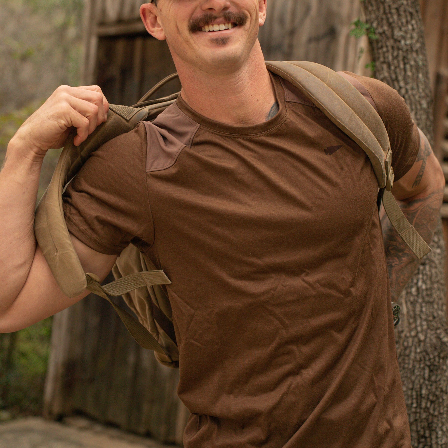 A smiling man wearing the Men’s Commando Tee - Merino Wool puts on a tan backpack outdoors beside a wooden building.