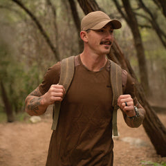 Man wearing tan cap and brown shirt with rucksack hiking through wooded trail