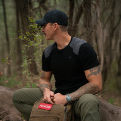 Man in black GORUCK shirt and cap sitting on log in forest with tactical rucking backpack