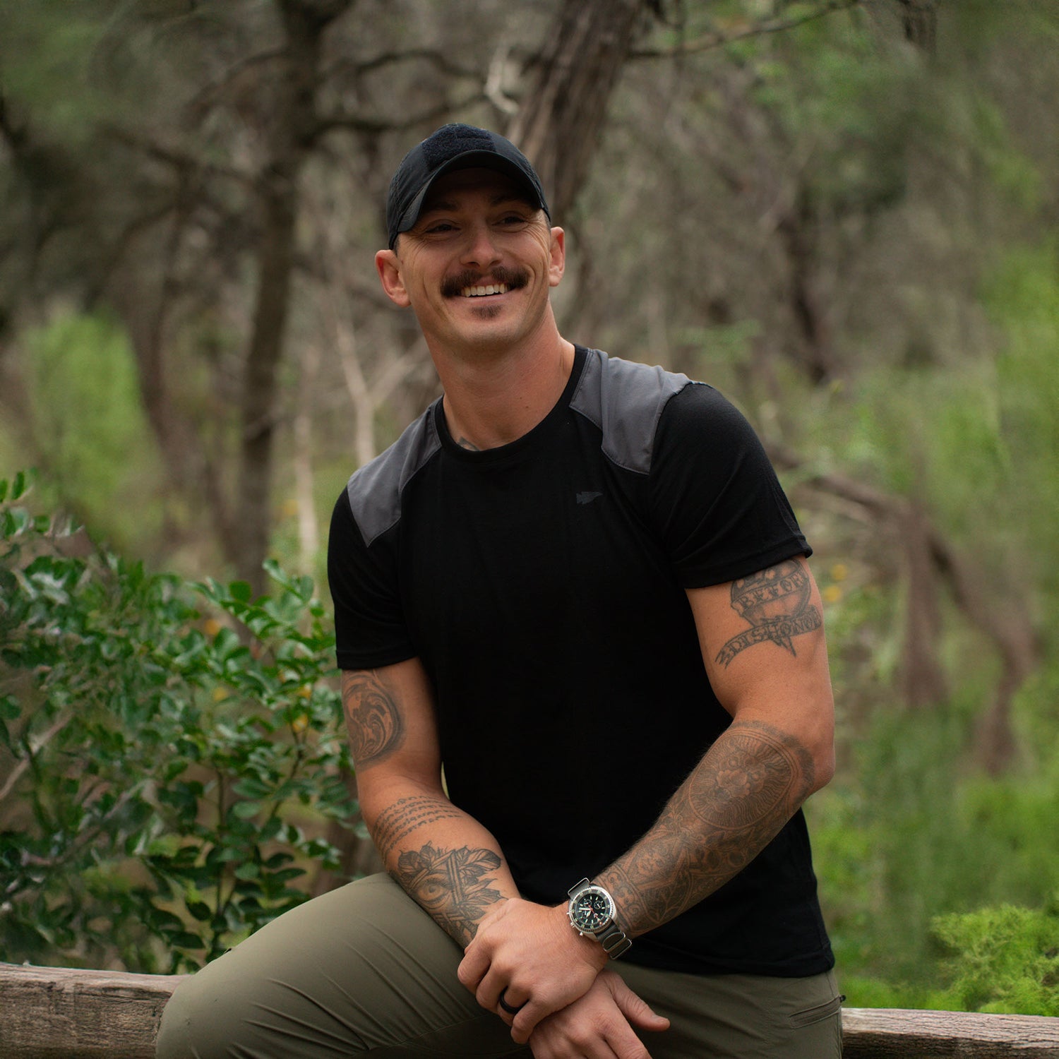 A tattooed man with a mustache smiles outdoors, surrounded by greenery, wearing the black Men’s Commando Tee - Merino Wool and a matching cap.