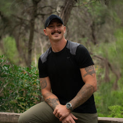 Smiling man in black GORUCK shirt and cap with tattooed arms in forest setting