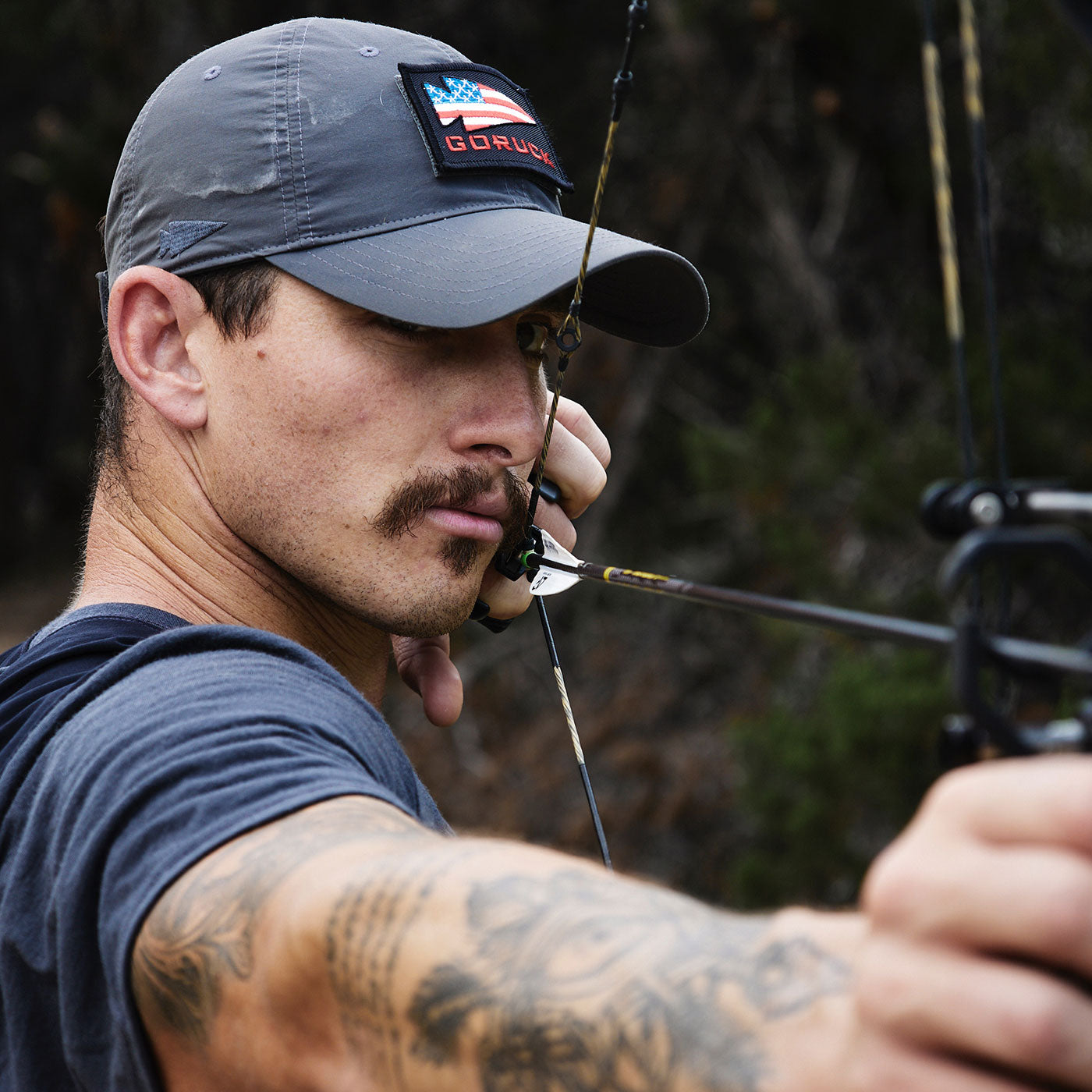 Man with a mustache aiming a compound bow, wearing a gray cap and a tattooed arm in focus.