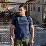 Man wearing GORUCK navy camo t-shirt and rucksack standing by a chain-link fence outdoors