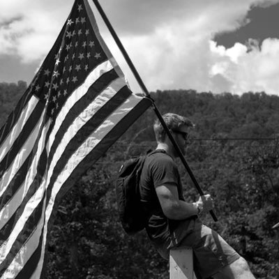 A man confidently sits on a wooden post, holding a large American flag. Wearing sunglasses and the durable Bullet Ruck Double Compartment - Dyneema backpack by GORUCK, he is surrounded by trees and hills under a partly cloudy sky. This striking black-and-white image captures a timeless moment of strength and resilience.